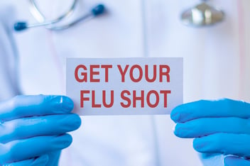 Person in lab coat wearing blue gloves and holding a sign that says: Get your flu shot