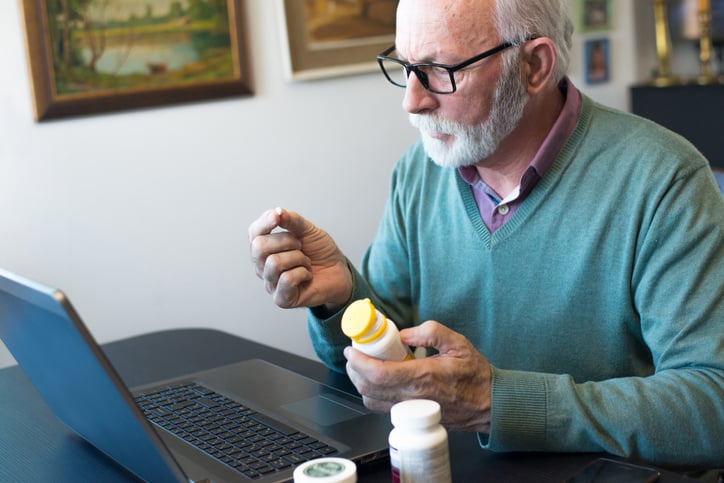Older man looking at medication and referring to information on a laptop.
