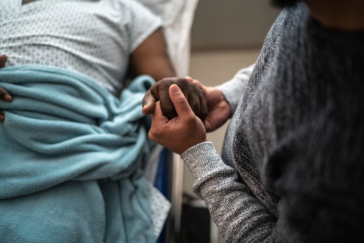 Black male patient in hospital bed, holding a loved one's hand.