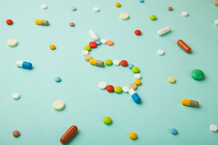 Colorful pills and capsules form a dollar sign on a light blue background.